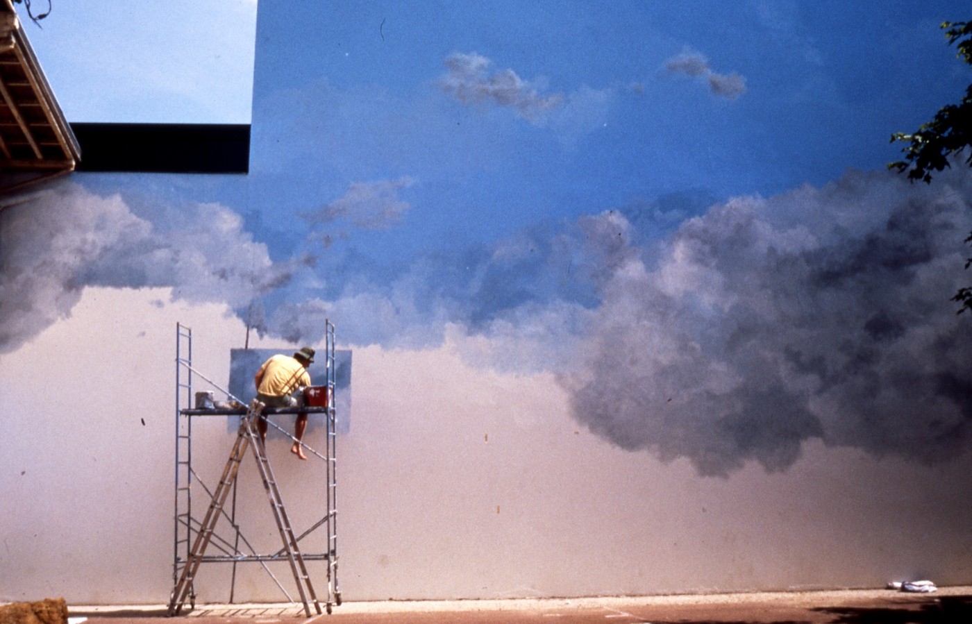 Peintre sur échafaudage en train de peindre un ciel et des nuages en grand format.