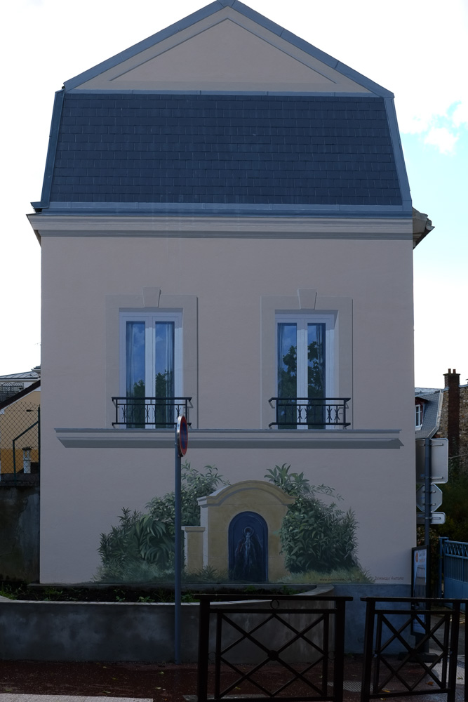 Vu de l’autre côté de la rue où des trains qui passent devant, le trompe-l’oeil prend part discrètement au paysage urbain. À Castelnau-le-Lez, à côté de Montpellier, une glycine. Trompe-l’oeil architectural et pictural